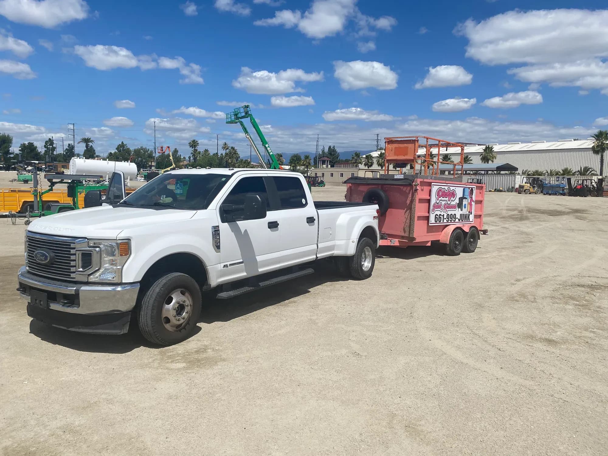A white pickup truck attached to Pinky's bright pink junk-removal trailer at a commercial yard.